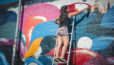 Photo d'une femme sur une échelle appuyée contre un mur en train de tracer un trait avec une bombe de peinture